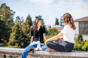 Two people sitting outside Doe library, facing each other. One looks to the side.